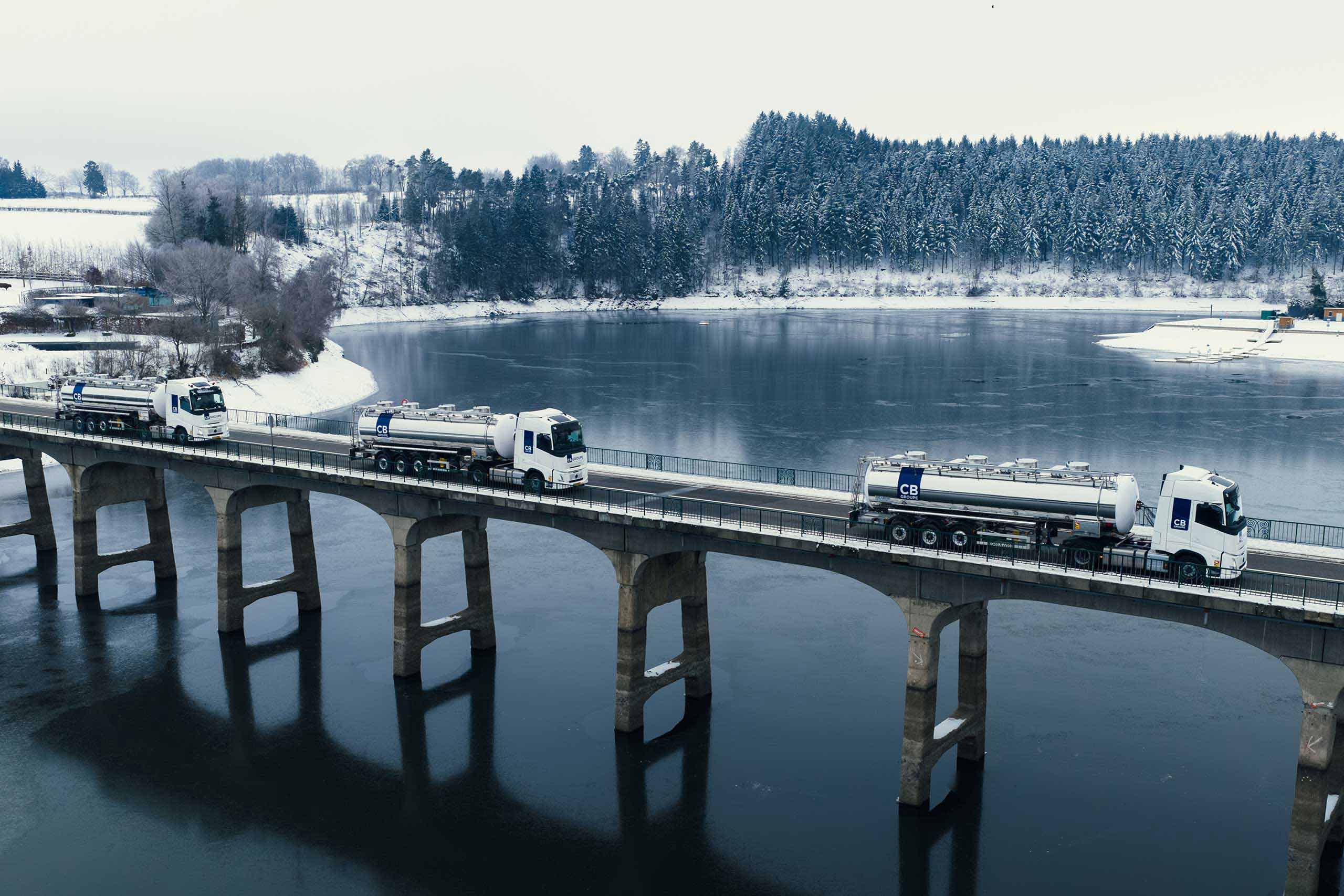 Plusieurs camions-citernes blancs sur un pont au-dessus d’un lac gelé, entouré de neige.