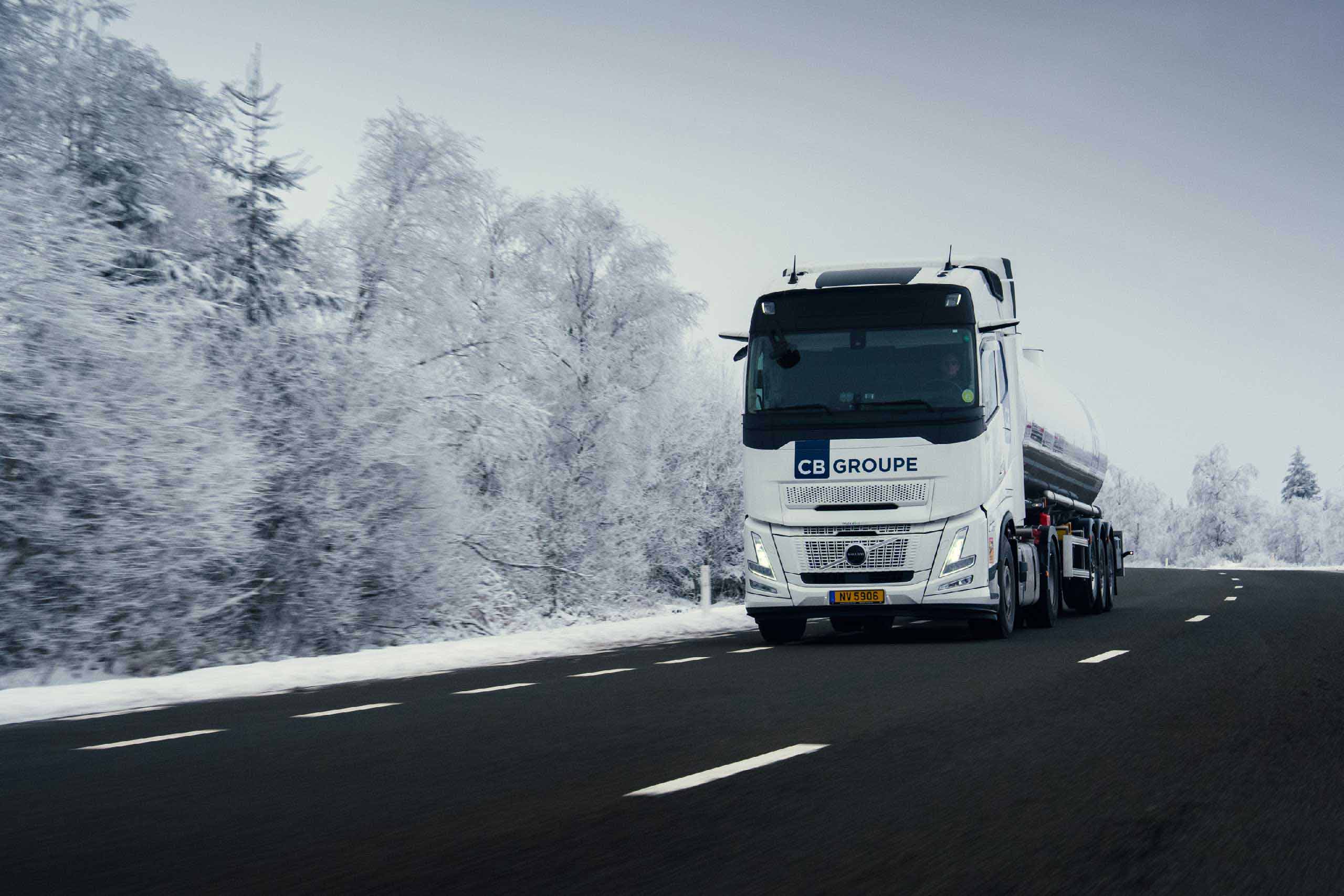 Camion-citerne blanc CB GROUPE roulant sur une route enneigée bordée d’arbres givrés.