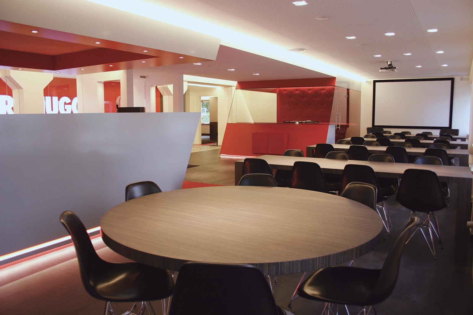 Un espace de réunion moderne avec des tables rondes et des chaises noires. Des murs blancs et rouges, avec un écran de projection sur le mur. L'éclairage est doux et le sol est de couleur sombre.
